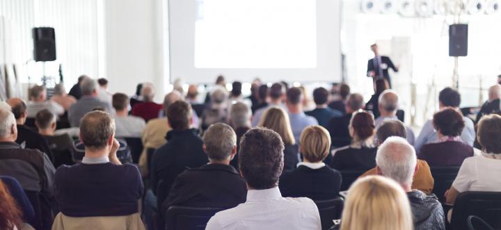 View of audience from rear of room listening to a suited male present.