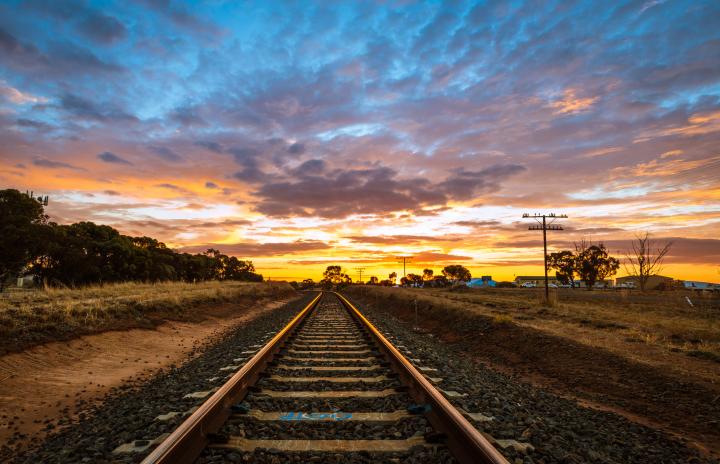 An image of railway tracks in a country landscape running towards a horizon with the sun setting in the background.