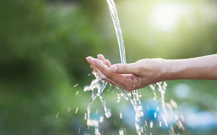 An image of an outstretched hand with water pouring into it, in an outdoor setting. 