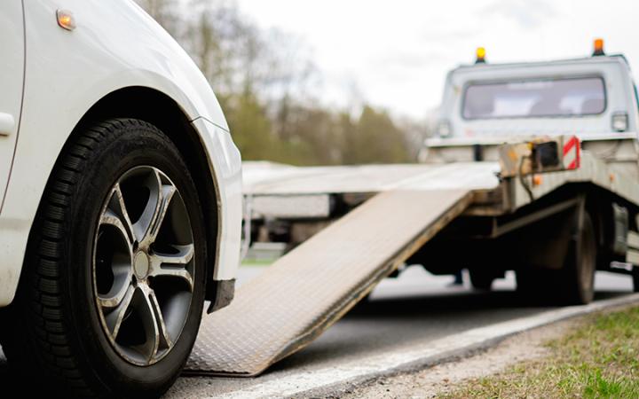 A tow truck in the process of rolling a broken-down car up ramps on to the truck's tray. 