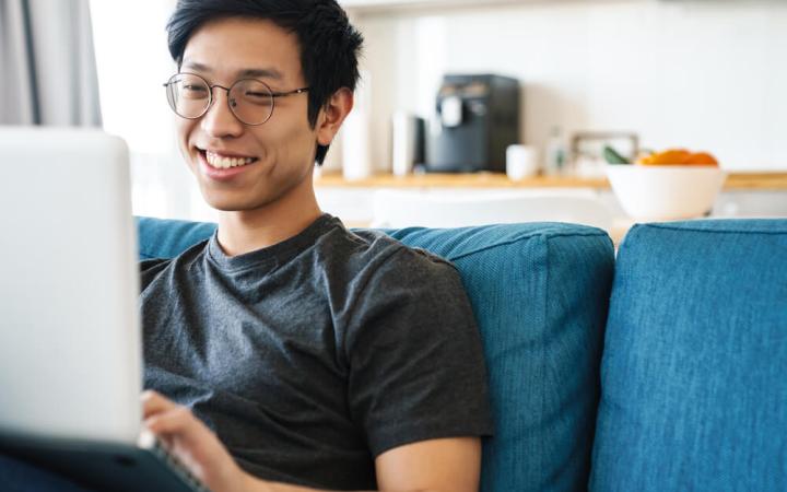 A photo of a man sitting on a couch and smiling while using a laptop.
